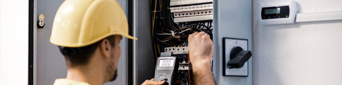 an electrical wearing a yellow hardhat checking voltage on an electrical panel enclosure